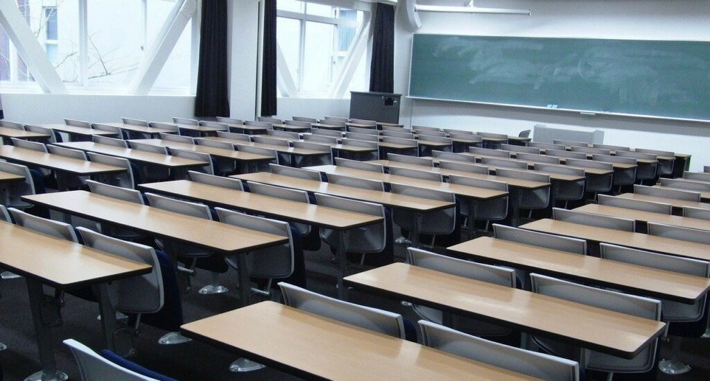 Aula de universidad, con pupitres, mesa de profesor y pizarra sin texto.