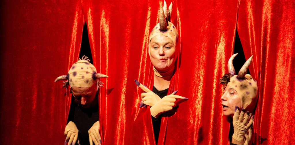 Fotografía de una obra de teatro accesible, con tres personas disfrazadas de animales con un telón rojo