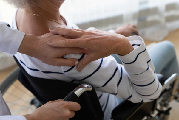 Mujer mayor en silla de ruedas, de espaldas, dando la mano a su cuidador.