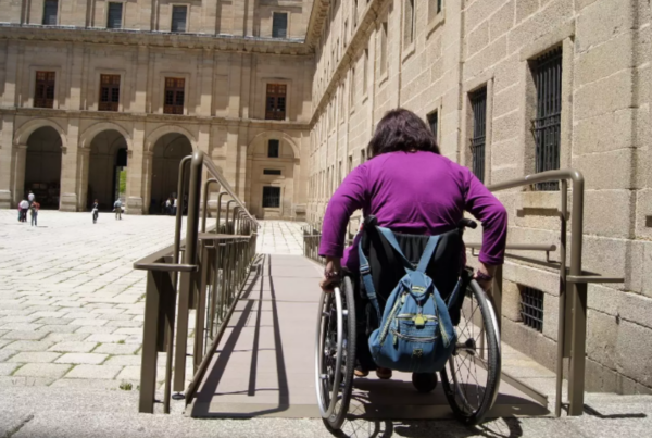 Una mujer joven en silla de ruedas, de espaldas, desciende por una rampa en una plaza monumental de un destino turístico