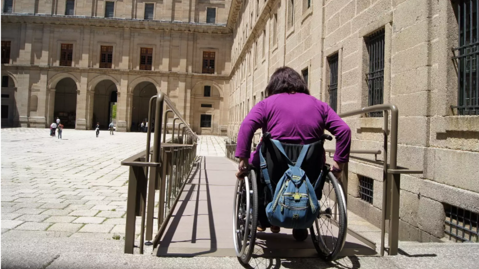 Una mujer joven en silla de ruedas, de espaldas, desciende por una rampa en una plaza monumental de un destino turístico