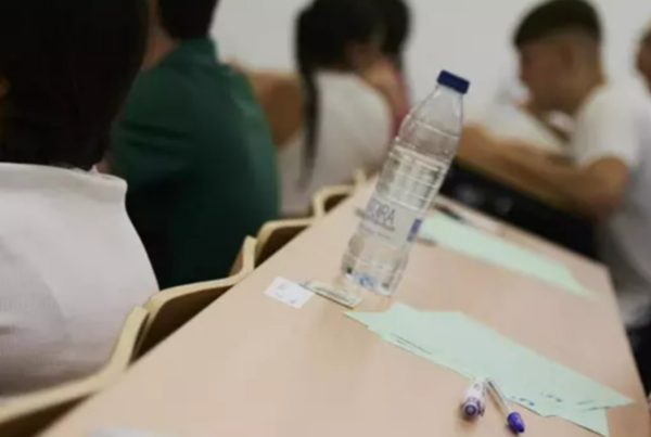 Fotografía del aula de una universidad, con una mesa con papeles y bolígrafos y varios alumnos, con imagen difuminada.