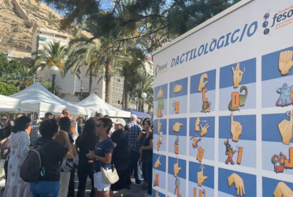 Fotografía de la jornada organizada por Fesord en Alicante por el Día Internacional de las Personas Sordas. A la izquierda, se ven grupos de personas hablando con lengua de signos y, al fondo, los stands. A la derecha, un mural con el abecedario en lengua de singos: Dactilológico