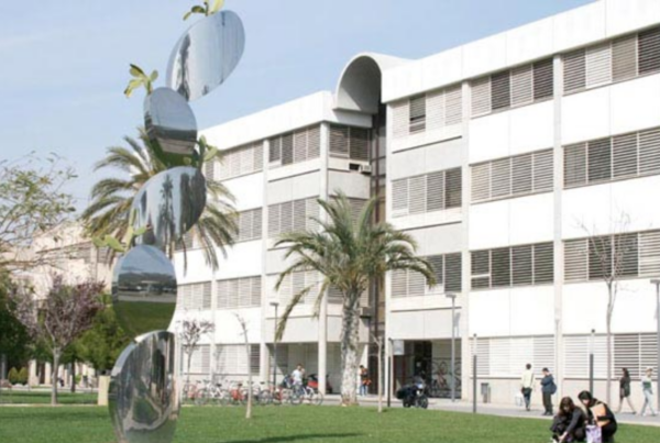 Fotografía del campus de la UPV, con una de las facultades, una zona de césped delante y algunas personas sentadas en el mismo