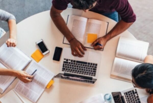 tres estudiantes trabajando en una mesa redonda, con libros, móviles y portátiles