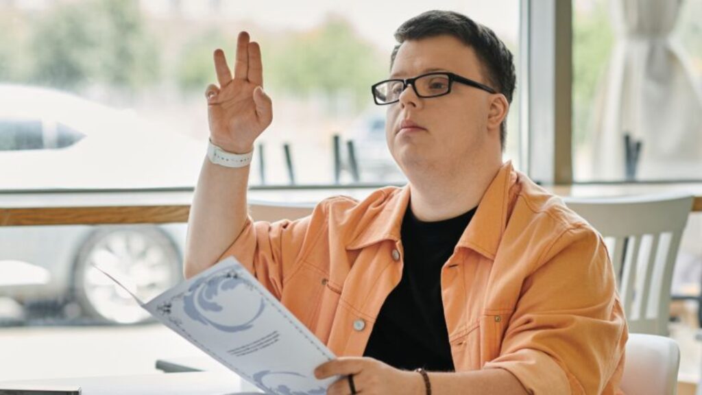 fotografía de un joven con síndrome de down, sentado en una mesa de un restaurante. Tiene la carta de productos en la mano y está levantando la mano para llamar al personal.