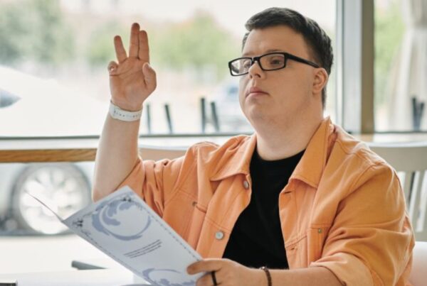 fotografía de un joven con síndrome de down, sentado en una mesa de un restaurante. Tiene la carta de productos en la mano y está levantando la mano para llamar al personal.