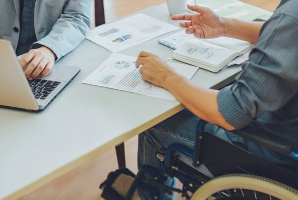 un hombre joven en silla de ruedas conversa con un compañero de trabajo, mientras revisan documentos en papel y en la pantalla de un ordenador portátil