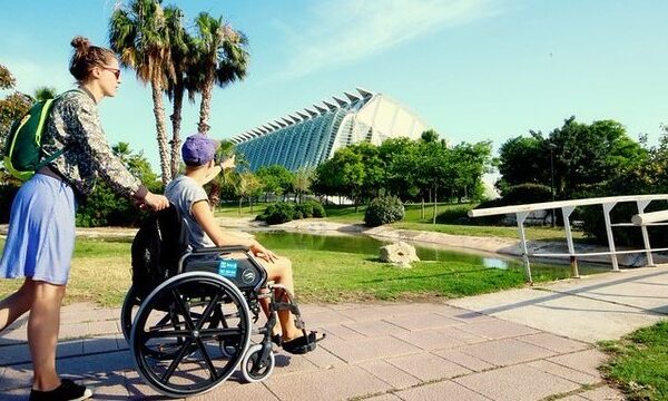 fotografía de una pareja de turistas jóvenes, chico y chica, paseando en el parque del río Turia, en el entorno del Museo Príncipe Felipe. El chico va en silla de ruedas.