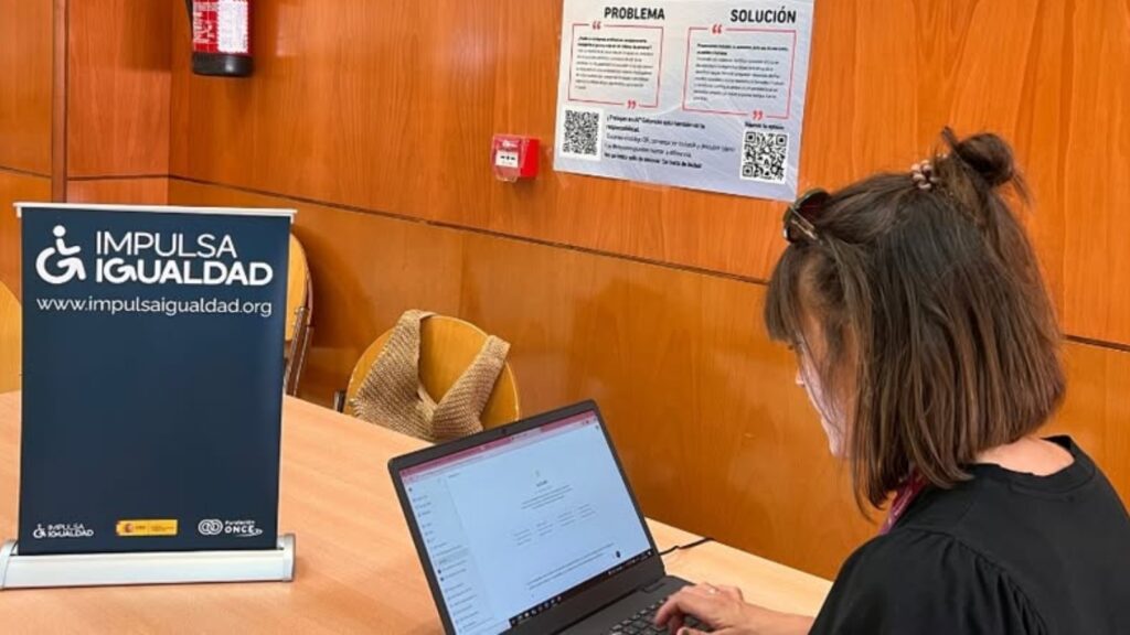 fotografía de una mujer joven trabajando con su ordenador portátil. Sobre la mesa, hay un cartel de Impulsa Igualdad