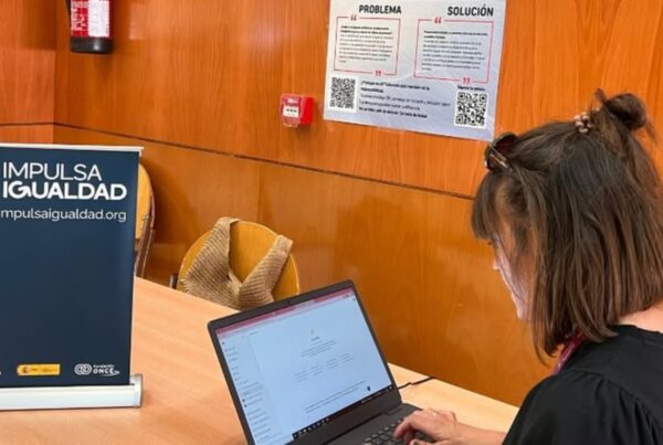 fotografía de una mujer joven trabajando con su ordenador portátil. Sobre la mesa, hay un cartel de Impulsa Igualdad