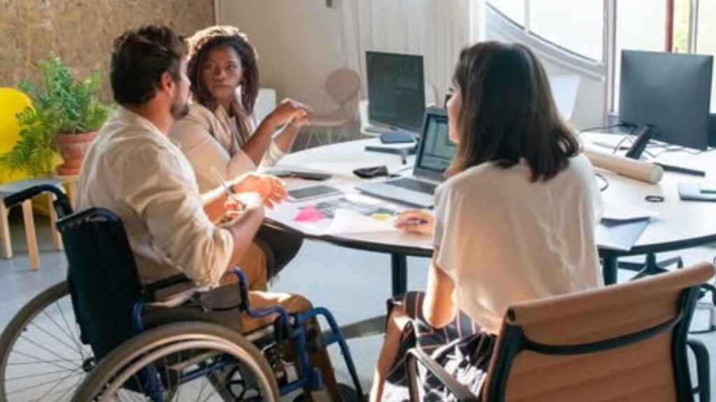 fotografía de un grupo de trabajo en una empresa, formado por tres personas jóvenes, dos mujeres y un hombre, que está en una silla de ruedas. Están debatiendo alrededor de un ordenador portátil.