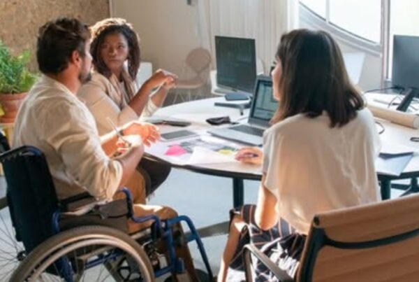 fotografía de un grupo de trabajo en una empresa, formado por tres personas jóvenes, dos mujeres y un hombre, que está en una silla de ruedas. Están debatiendo alrededor de un ordenador portátil.