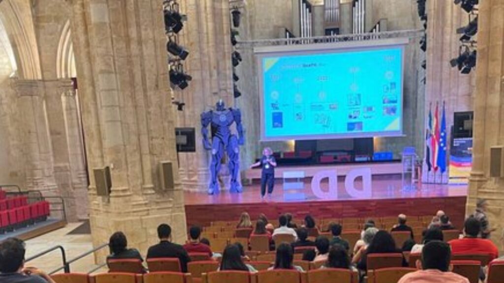 Fotografía de chele esteve durante su intervención. Es una panorámica general de la sala, en la que se observan varios asistentes y, al fondo, el escenario general. Chele está arriba y en la pantalla está proyectada información sobre la evolución de dicatic