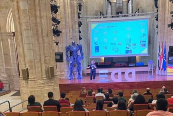 Fotografía de chele esteve durante su intervención. Es una panorámica general de la sala, en la que se observan varios asistentes y, al fondo, el escenario general. Chele está arriba y en la pantalla está proyectada información sobre la evolución de dicatic