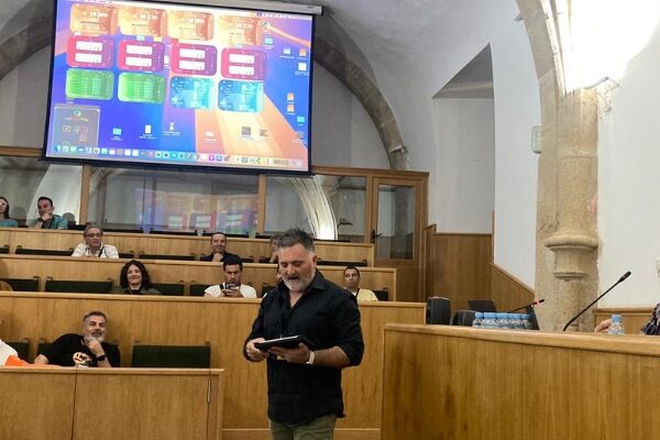 fotografía del taller. El profesor está hablando al tiempo que muestra en una gran pantalla las aplicaciones desarrolladas. Las personas participantes están sentadas en una especie de hemiciclo.