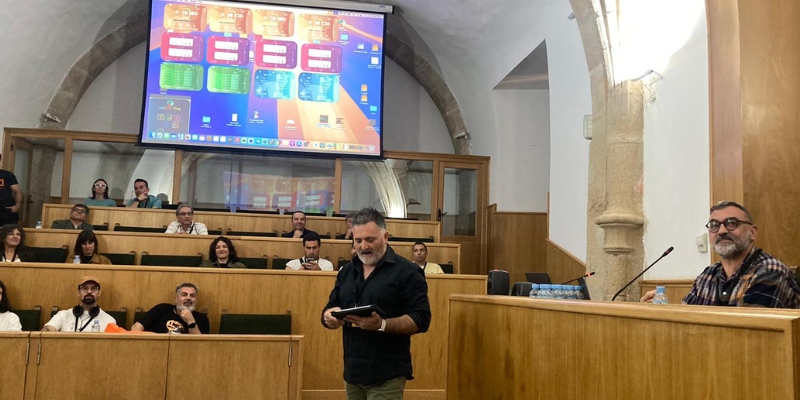 fotografía del taller. El profesor está hablando al tiempo que muestra en una gran pantalla las aplicaciones desarrolladas. Las personas participantes están sentadas en una especie de hemiciclo.