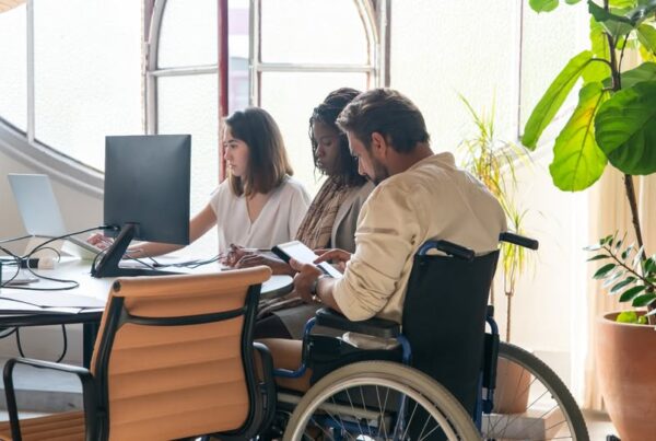 Equipo de tres personas, una de ellas en silla de ruedas, en torno a una mesa de escritorio y trabajando con un ordenador. El hombre en silla de ruedas está consultando su tablet.