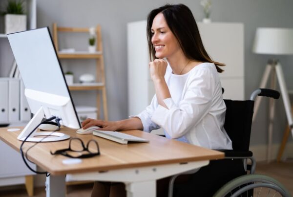 una mujer joven en silla de ruedas está sentada frente a una mesa trabajando con su ordenador