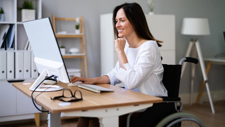 una mujer joven en silla de ruedas está sentada frente a una mesa trabajando con su ordenador