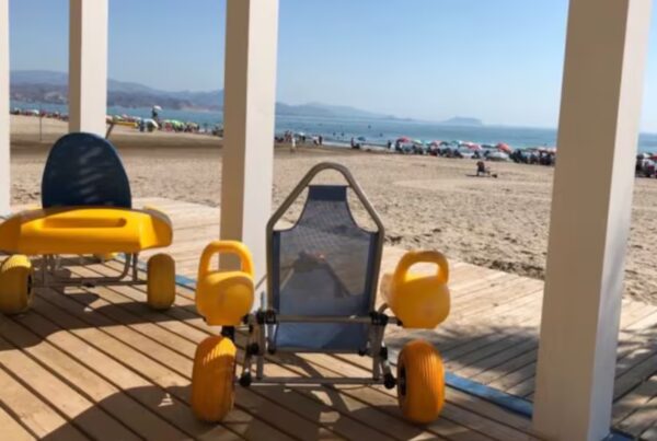 fotografía de dos sillas de ruedas adaptadas situadas sobre una pasarela de madera de una playa. Al fondo se ve la arena y el mar.