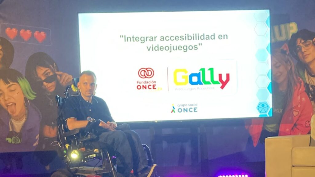 fotografía de cristian durante su ponencia en el congreso. Está encima del escenario, en su silla de ruedas. Detrás, en la pantalla, aparece el título de su ponencia: integrar accesibilidad en videojuegos