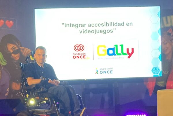 fotografía de cristian durante su ponencia en el congreso. Está encima del escenario, en su silla de ruedas. Detrás, en la pantalla, aparece el título de su ponencia: integrar accesibilidad en videojuegos
