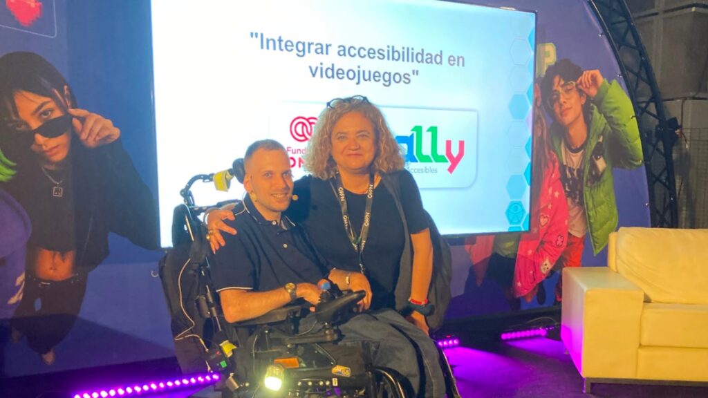 Fotografía de cristian y chele esteve en el escenario del evento. De fondo, la pantalla con el título de la ponencia: integrar accesibilidad en videojuegos